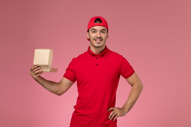 Vista frontal do mensageiro masculino com uniforme vermelho e boné segurando um pequeno pacote de entrega e sorrindo na parede rosa