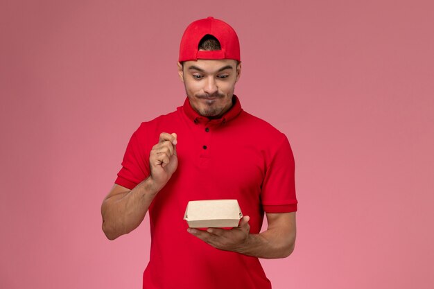 Vista frontal do mensageiro masculino de uniforme vermelho e boné segurando um pequeno pacote de entrega na parede rosa