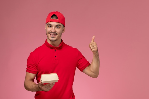 Vista frontal do mensageiro masculino de uniforme vermelho e boné segurando um pequeno pacote de entrega na parede rosa