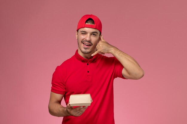 Vista frontal do mensageiro masculino de uniforme vermelho e boné segurando um pequeno pacote de entrega na parede rosa