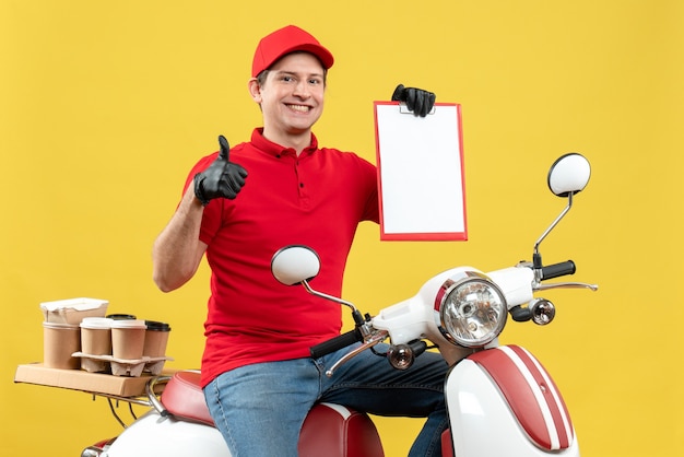 Vista frontal do mensageiro usando blusa vermelha e luvas de chapéu na máscara médica, entregando o pedido, sentado na scooter, segurando o documento, fazendo um gesto de ok