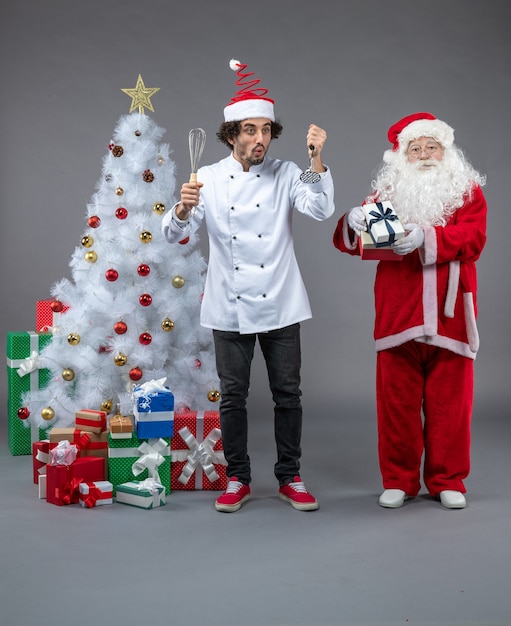 Vista frontal do Papai Noel com cozinheiro masculino em volta dos presentes de Natal na parede cinza