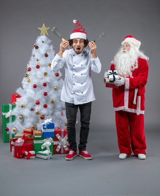 Vista frontal do Papai Noel com cozinheiro masculino em volta dos presentes de Natal na parede cinza