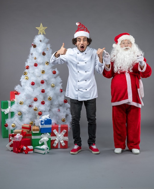 Foto grátis vista frontal do papai noel com cozinheiro masculino em volta dos presentes de natal na parede cinza