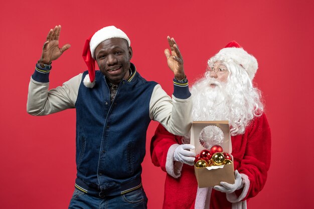 Vista frontal do Papai Noel com jovem segurando brinquedos da árvore de natal na parede vermelha