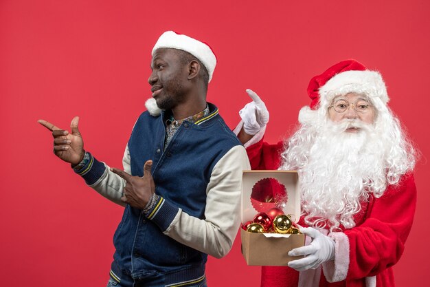 Vista frontal do Papai Noel com jovem segurando brinquedos da árvore de natal na parede vermelha