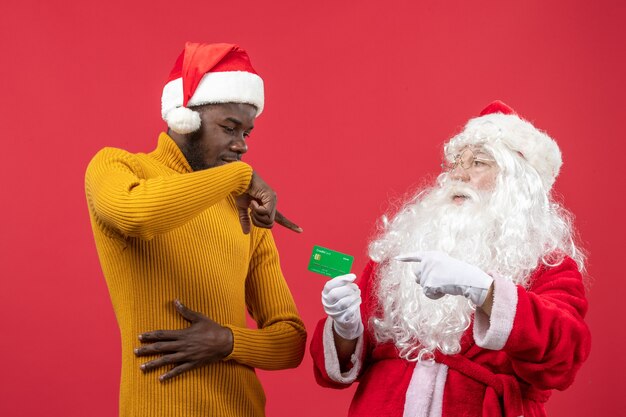 Vista frontal do Papai Noel com jovem segurando o cartão do banco em uma parede vermelha