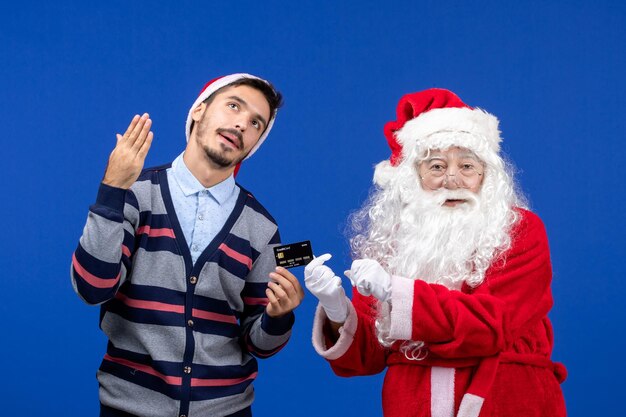 Vista frontal do papai noel com jovem segurando o cartão do banco na parede azul