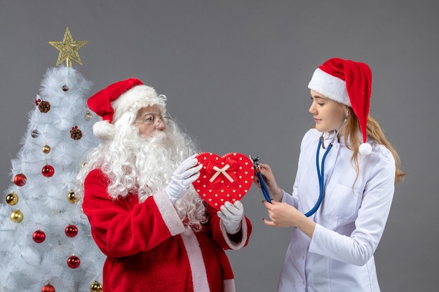 Vista frontal do Papai Noel com médica observando com estetoscópio na parede cinza