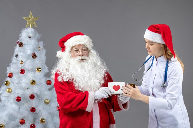 Vista frontal do Papai Noel com médica segurando o frasco com comprimidos na parede cinza