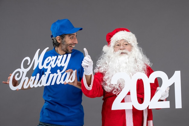 Vista frontal do papai noel com mensageiro segurando o feliz natal e as placas de 2021 na parede cinza