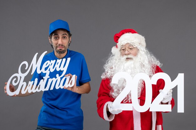 Foto grátis vista frontal do papai noel com mensageiro segurando o feliz natal e as placas de 2021 na parede cinza