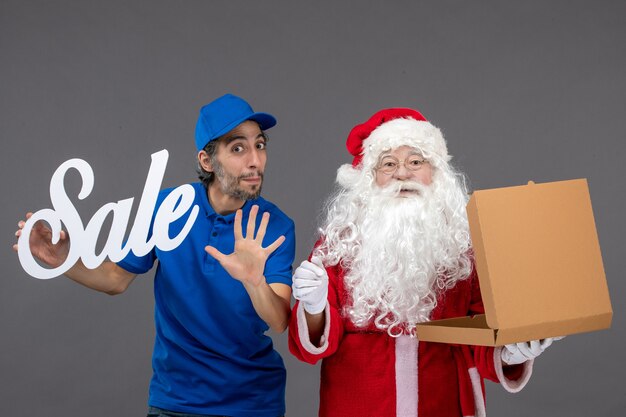 Vista frontal do Papai Noel com o correio masculino segurando a escrita de venda e a caixa de comida na parede cinza