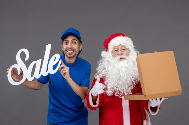 Vista frontal do papai noel com o mensageiro masculino segurando a faixa de venda e abrindo a caixa de comida na parede cinza