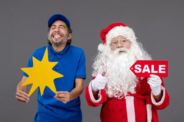 Vista frontal do papai noel com o mensageiro masculino segurando a faixa de venda e uma placa amarela na parede cinza