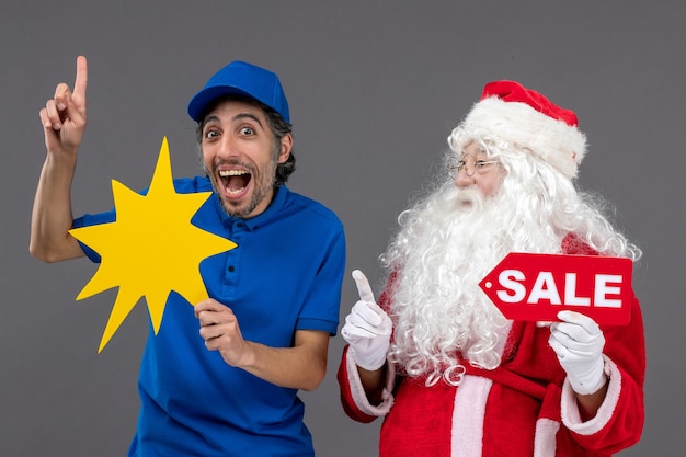 Foto grátis vista frontal do papai noel com o mensageiro masculino segurando a faixa de venda e uma placa amarela na parede cinza