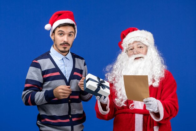 Vista frontal do Papai Noel com um jovem lendo carta e segurando um presente na parede azul