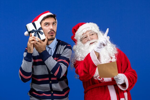 Vista frontal do Papai Noel com um jovem lendo carta e segurando um presente na parede azul