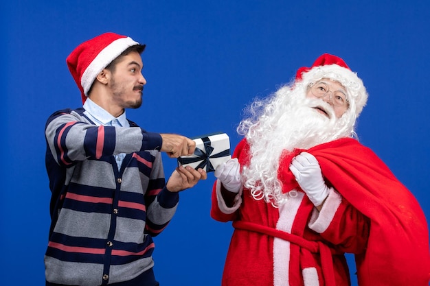 Foto grátis vista frontal do papai noel com um jovem segurando um presente na parede azul