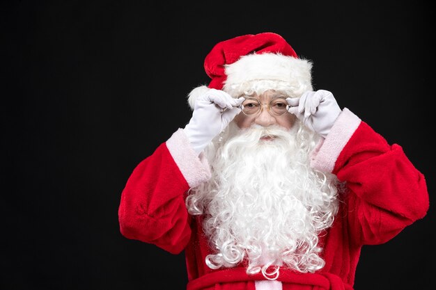Vista frontal do Papai Noel em um terno vermelho clássico com barba branca em pé na parede preta