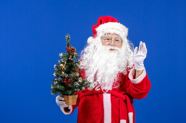 Vista frontal do papai noel segurando a pequena árvore de ano novo na neve azul ano novo natal