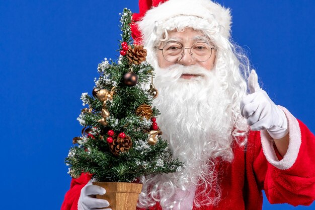 Vista frontal do papai noel segurando a pequena árvore de ano novo na neve azul colore o natal