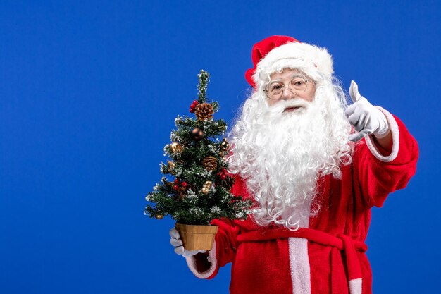 Vista frontal do papai noel segurando a pequena árvore de ano novo no chão azul, cor de Natal de ano novo