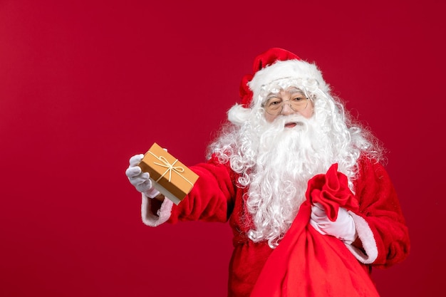 Vista frontal do papai noel segurando um presente de uma sacola cheia de presentes para as crianças na emoção vermelha do ano novo