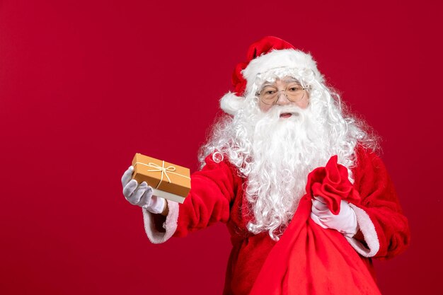 Vista frontal do papai noel segurando um presente de uma sacola cheia de presentes para as crianças na emoção vermelha do ano novo