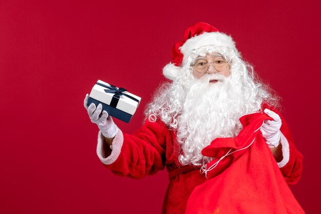 Vista frontal do papai noel segurando um presente de uma sacola cheia de presentes para as crianças no Natal de ano novo vermelho