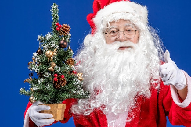 Vista frontal do papai noel segurando uma pequena árvore de ano novo em uma neve azul de natal