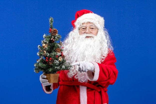 Vista frontal do papai noel segurando uma pequena árvore de ano novo na mesa azul de neve cor de natal