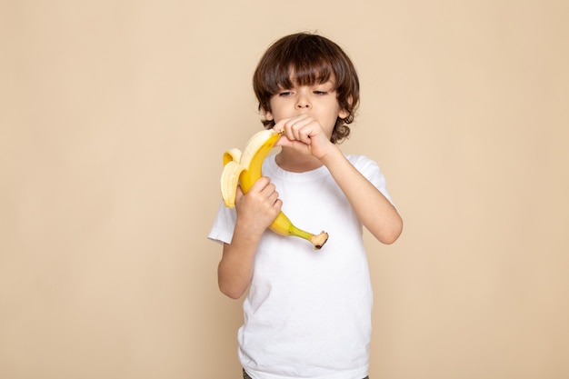vista frontal do retrato, garotinho fofo adorável em camiseta branca descascando bananon rosa