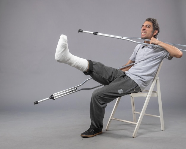 Vista frontal do sexo masculino jovem sentado com o pé quebrado usando muletas para andar na parede cinza acidente torção do pé dor na perna quebrada
