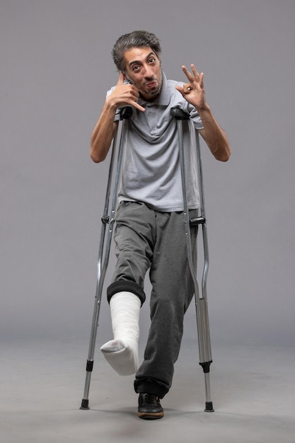 Vista frontal do sexo masculino jovem usando muletas devido a um pé quebrado na parede cinza Dor no pé incapacita a perna quebrada