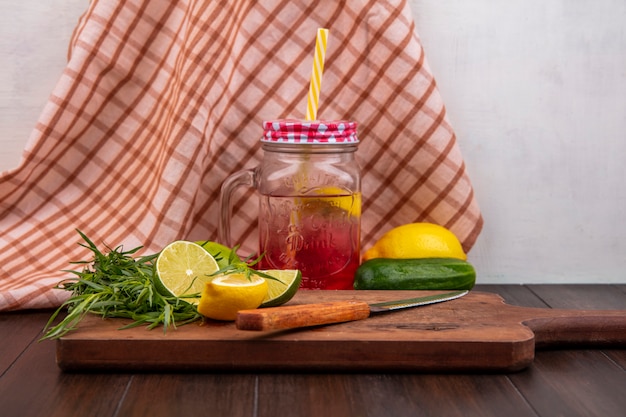Vista frontal do suco de limão fresco em uma jarra de vidro com limões verdes de estragão em uma placa de cozinha de madeira com faca em uma superfície de toalha de mesa marcada