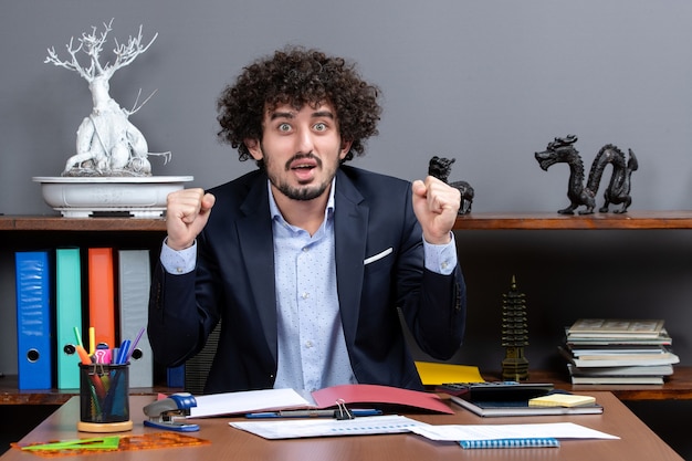 Vista frontal do trabalhador de escritório feliz mostrando gesto vencedor sentado à mesa no escritório