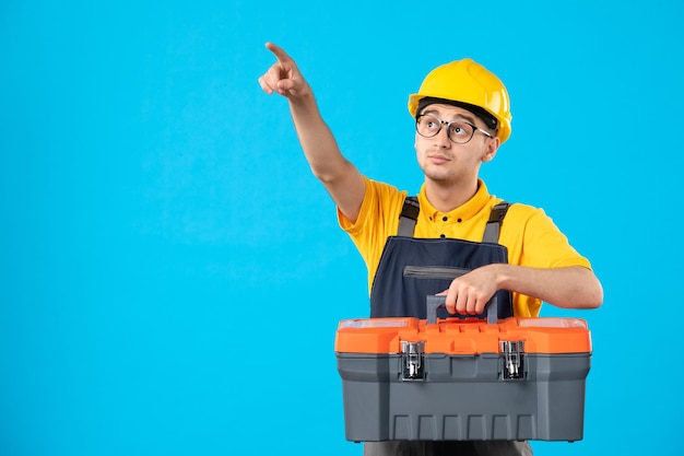 Vista frontal do trabalhador masculino em uniforme amarelo apontando para cima com caixa de ferramentas em azul