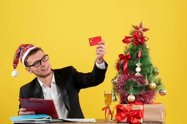 Vista frontal do trabalhador masculino segurando o cartão do banco e uma nota ao redor da pequena árvore de Natal e presentes em amarelo