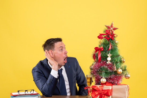 Vista frontal espantada com um homem ouvindo algo sentado à mesa perto da árvore de natal e presentes sobre fundo amarelo