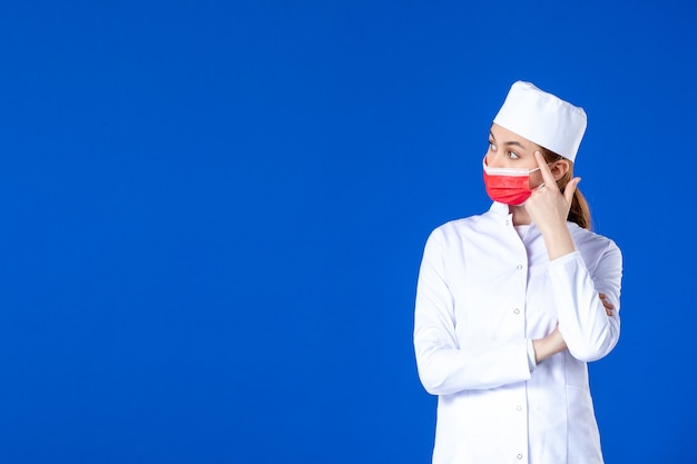 Vista frontal estressada jovem enfermeira em traje médico com máscara protetora vermelha na parede azul