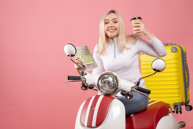 Vista frontal exultante jovem em ciclomotor segurando mapa e café elegante parede rosa