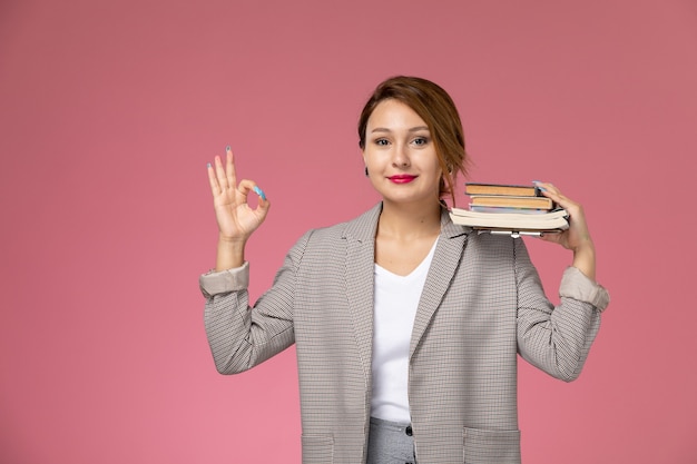 Vista frontal, jovem aluna com casaco cinza posando segurando livros e sorrindo no fundo rosa lições universidade estudo universitário