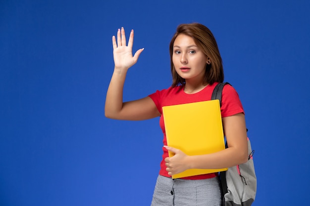 Vista frontal jovem aluna de camisa vermelha com mochila segurando arquivos amarelos acenando sobre fundo azul claro.