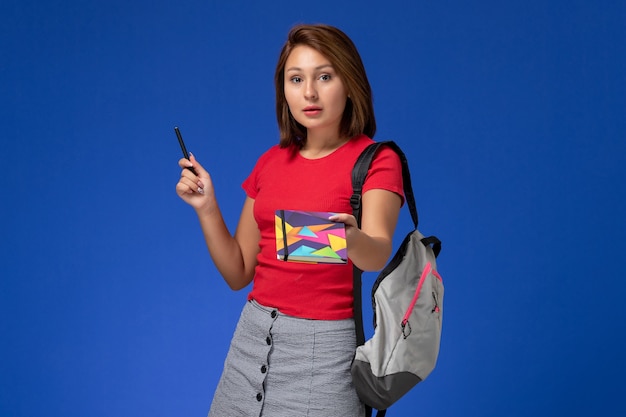 Vista frontal jovem aluna de camisa vermelha, usando mochila segurando a caneta com o caderno sobre o fundo azul claro.