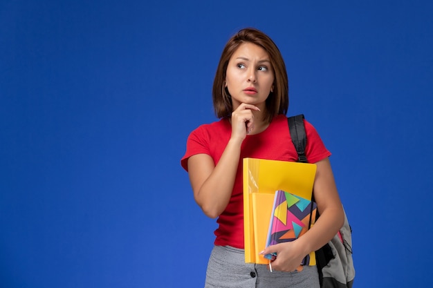 Foto grátis vista frontal jovem aluna de camisa vermelha, usando mochila segurando arquivos e caderno pensando sobre o fundo azul.