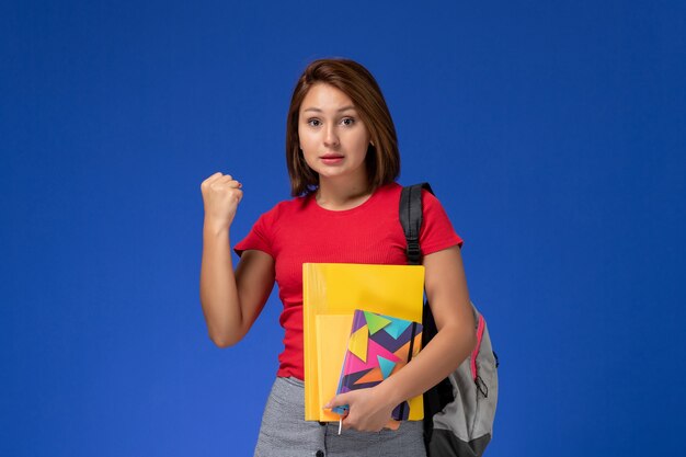 Vista frontal jovem aluna de camisa vermelha, usando mochila segurando arquivos e caderno regozijando-se sobre fundo azul.