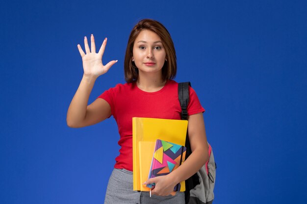 Vista frontal jovem aluna de camisa vermelha, usando mochila segurando arquivos e o caderno mostrando a palma da mão sobre fundo azul.