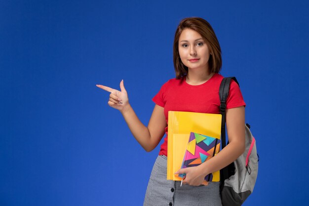 Vista frontal jovem aluna de camisa vermelha, usando mochila segurando arquivos e o caderno sobre o fundo azul.