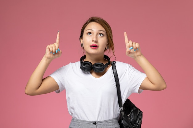 Vista frontal jovem aluna de camiseta branca e calça cinza com fones de ouvido olhando para o céu no fundo rosa.